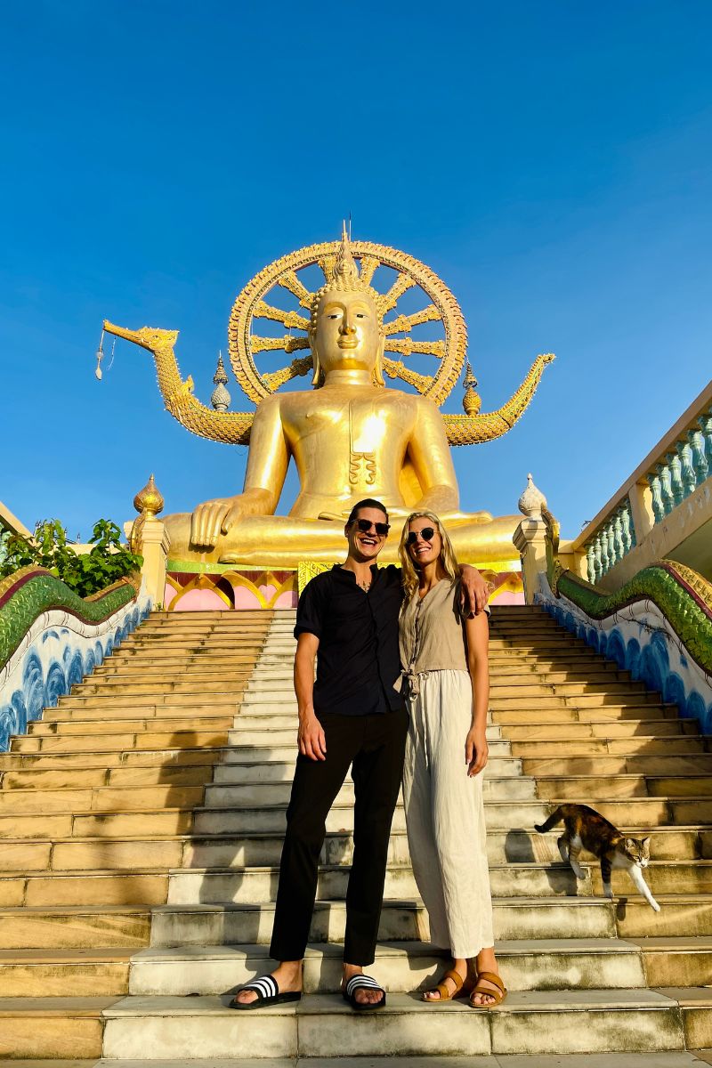 A couple standing on a set of stairs near a large golden Buddha statue in Koh Samui, with a dog and a cat sitting in front of them. In the background, there is a blue sky and a set of stairs leading to the statue.