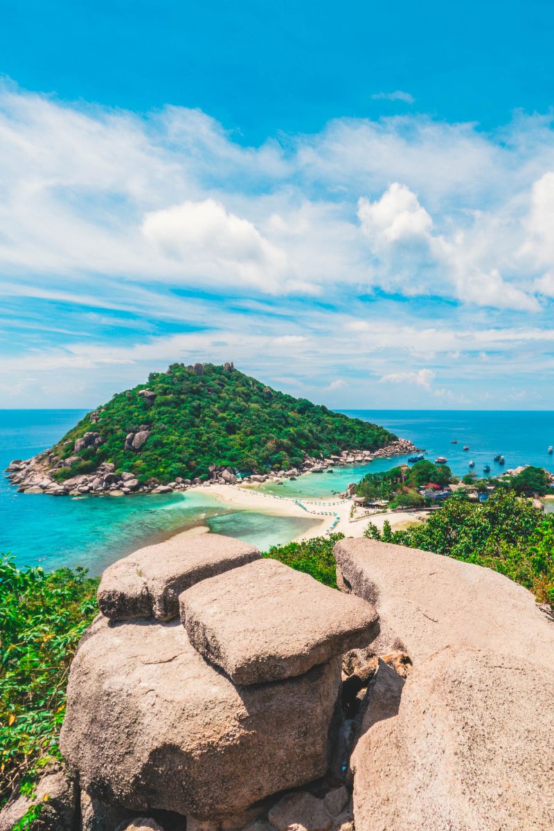 Island with lush greenery and clear blue water, viewed from a rocky outcrop.