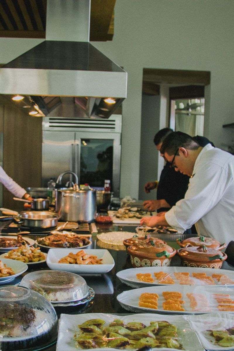 A buffet spread of prepared foods on trays and hot dishes on a stove in a kitchen or dining area, with several people serving themselves and others present in the scene.