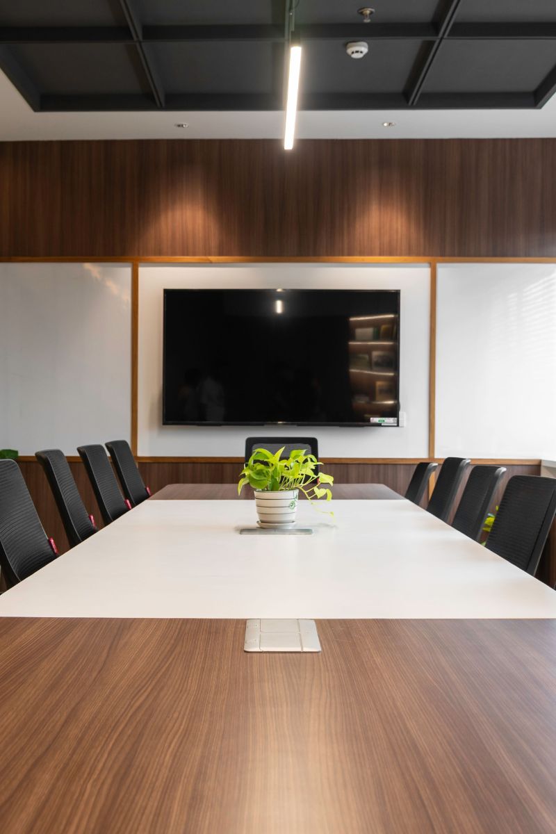 A modern conference room with a long rectangular table, black chairs, a wall-mounted TV, wood paneling, and a ceiling light fixture.