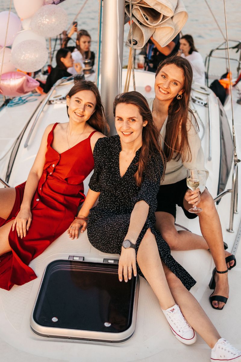 Three women posing together on the deck of a sailboat during what looks like a celebration, with balloons and other people visible in the background.