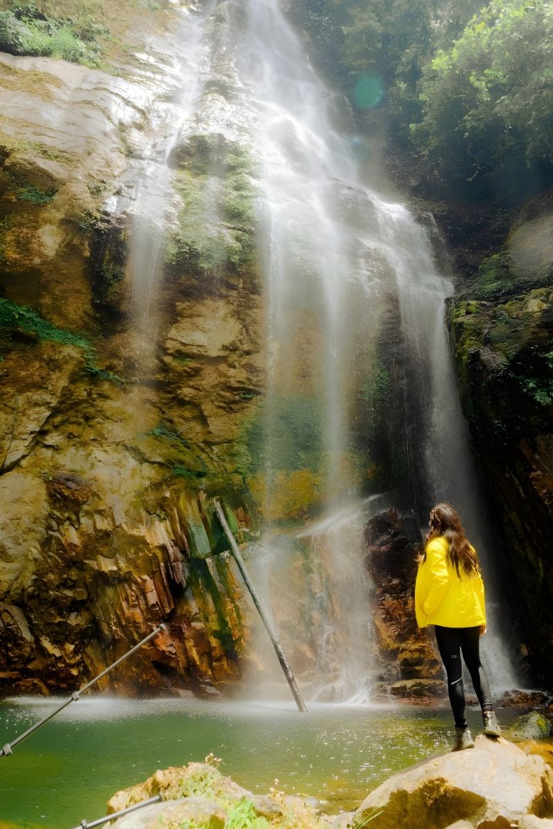 Person in a yellow jacket standing at the base of a waterfall in a forested area