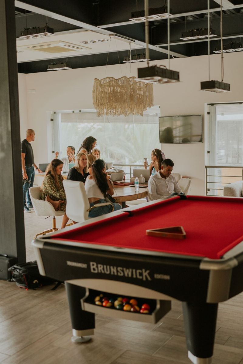 A Brunswick pool table with a red felt surface and black legs in a bright office or lounge space, with several people gathered in the background near a table and windows.