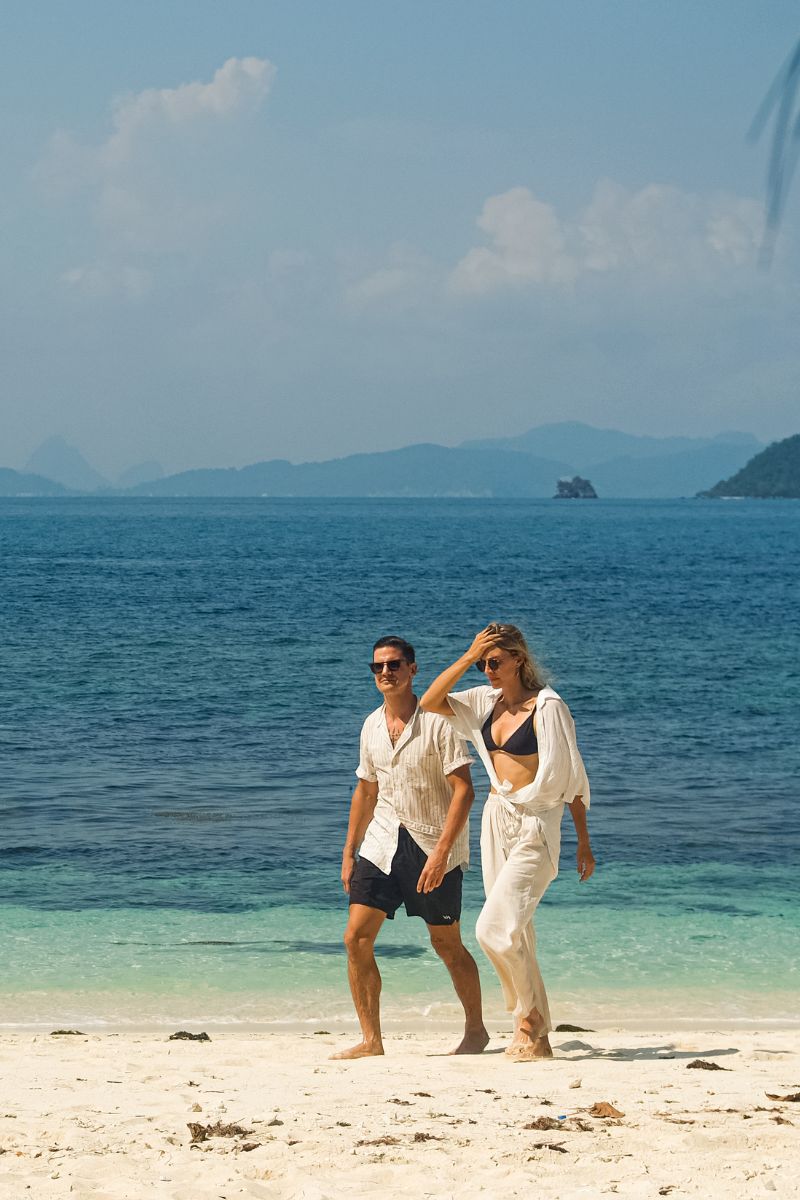 Two people walking on a beach with clear blue water and mountains in the background