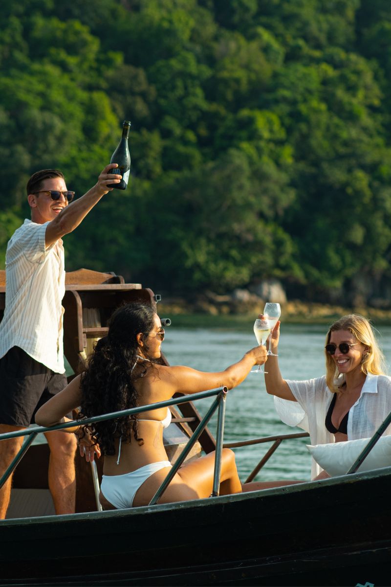 People on a boat with one person holding a bottle and another with a glass, surrounded by greenery.