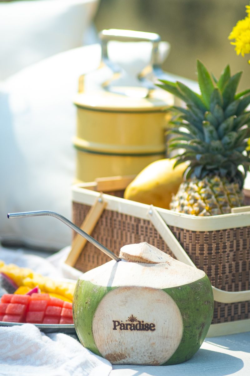 Coconut with a straw, pineapple, and fruit platter on a table outdoors.