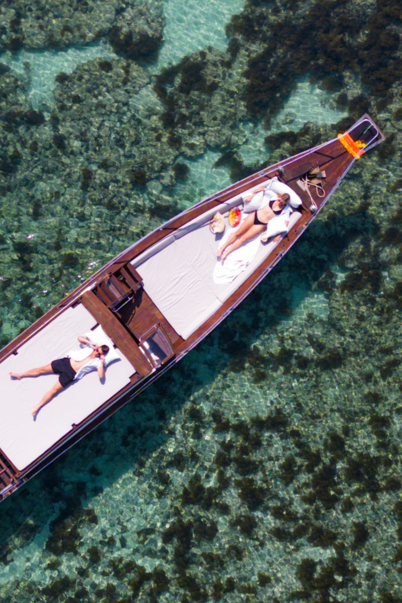 Aerial view of a longtail boat with people lounging on white towels in clear blue water.
