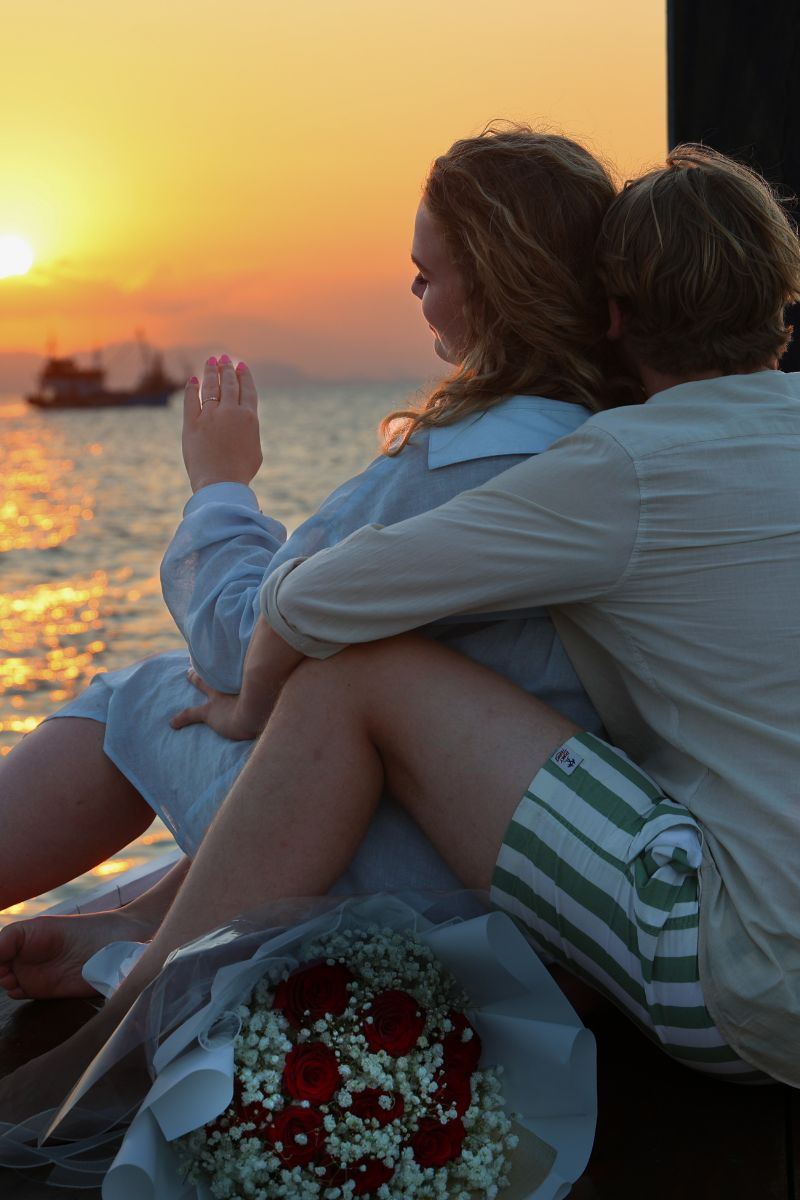 Couple sitting on a bench by the sea at sunset with a boat in the background.