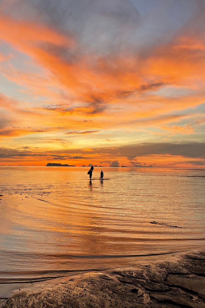 Two people walking on a beach at sunset with vibrant orange and purple clouds.