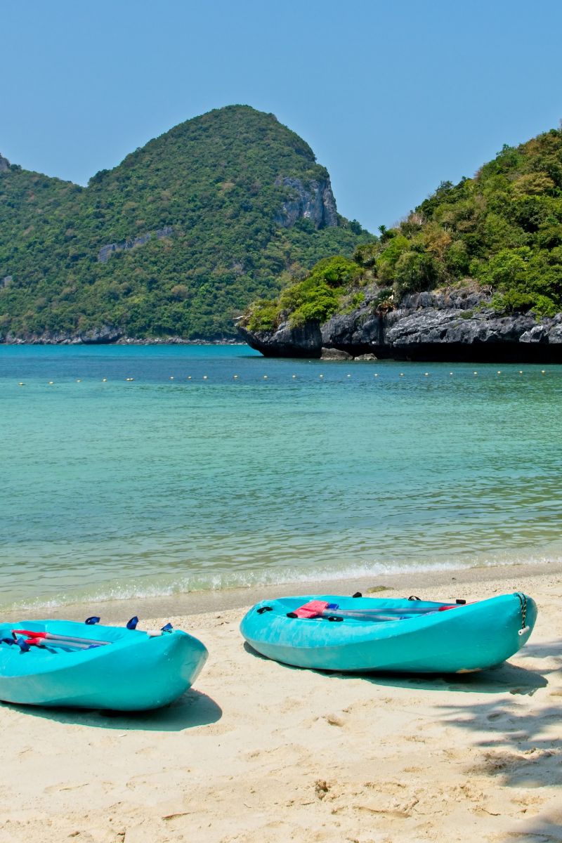 Two turquoise kayaks on a sandy beach with clear blue water and green mountains in the background.