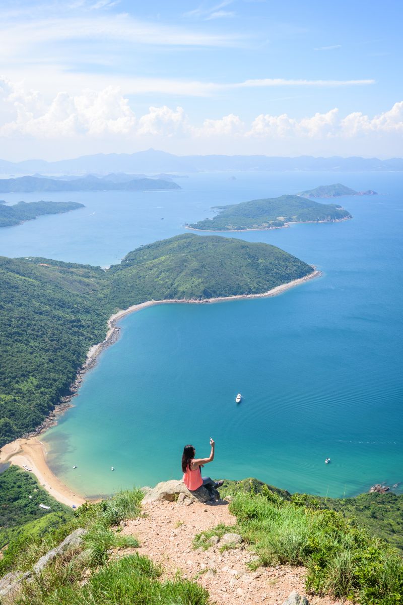 Person sitting on a cliff overlooking a scenic coastal view with green islands and blue water.
