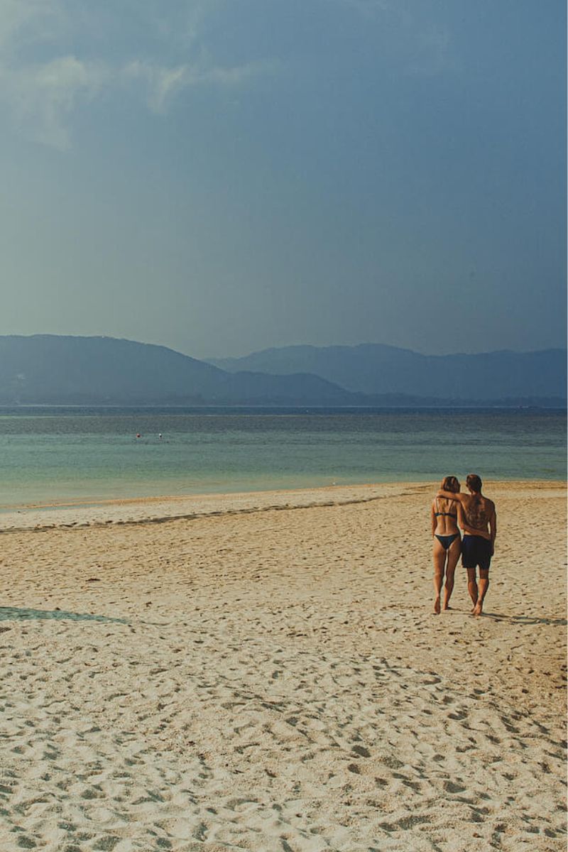 Two people walking on a sandy beach with mountains in the background