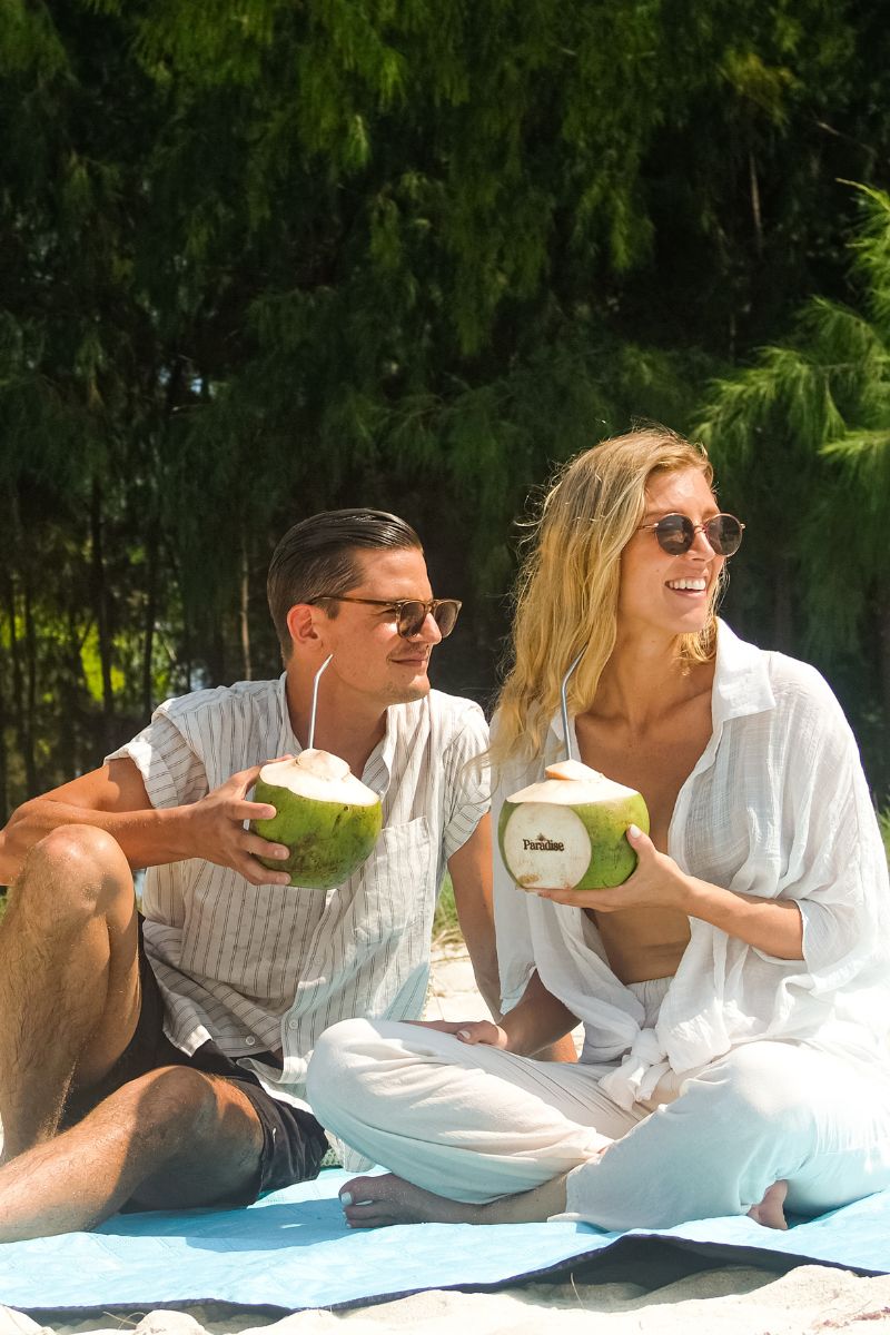 Two people sitting on a blanket outdoors, holding coconuts.