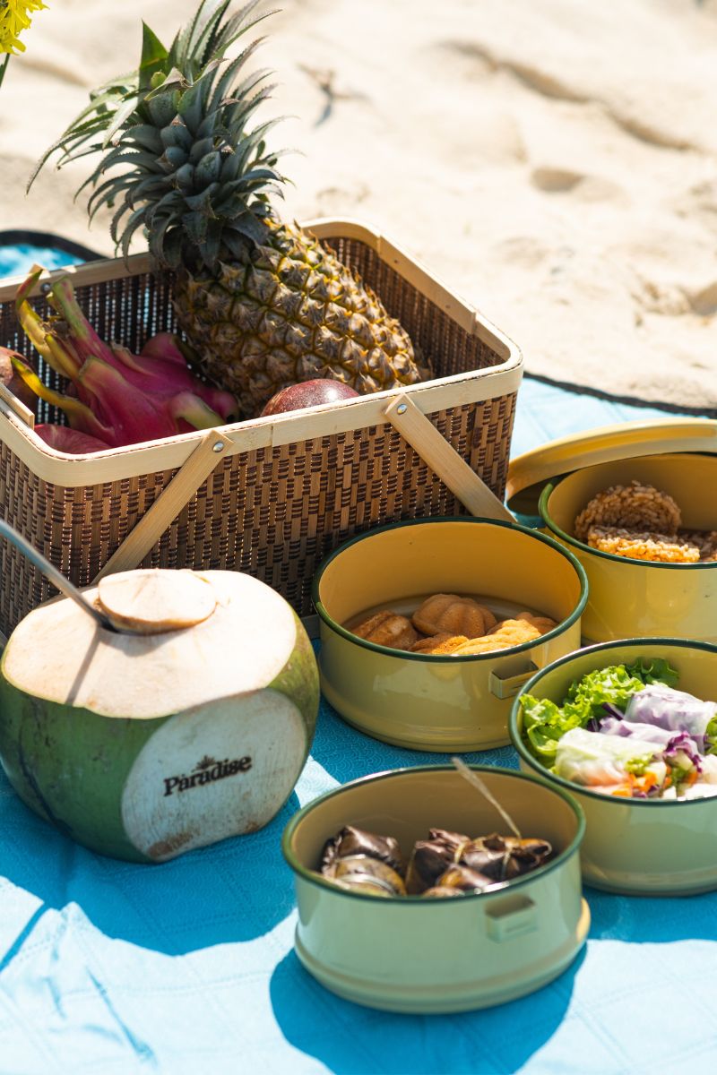 Picnic setup with a basket, coconuts, and various food items on a blue blanket.