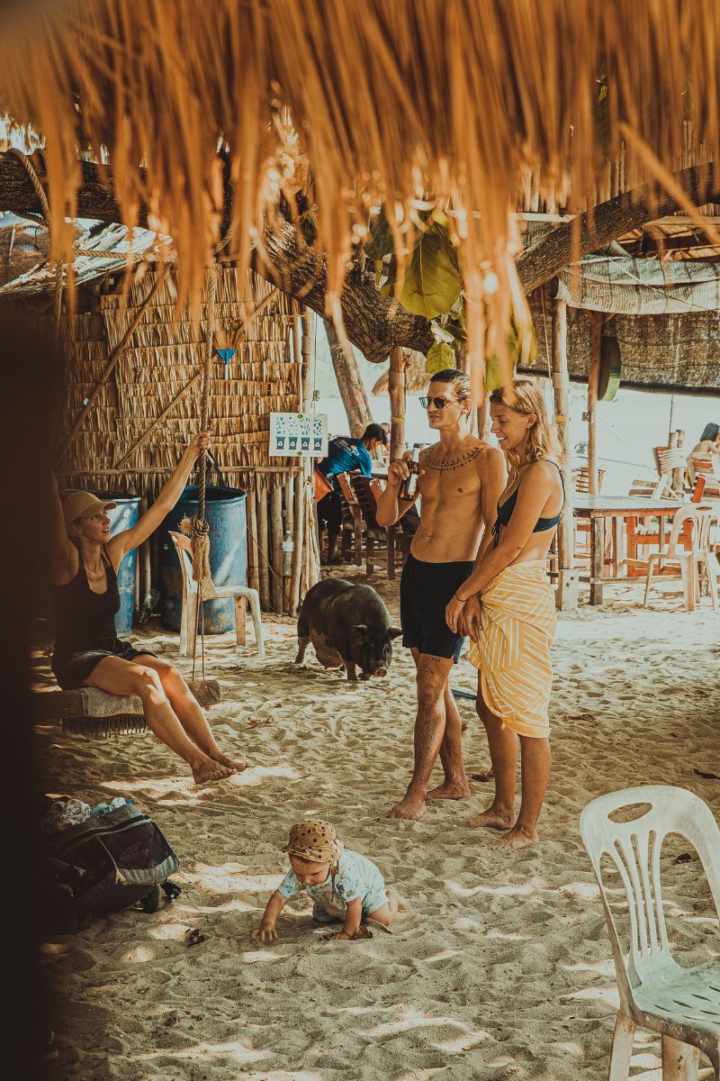 Family on a sandy beach with thatched-roof structures and people in the background