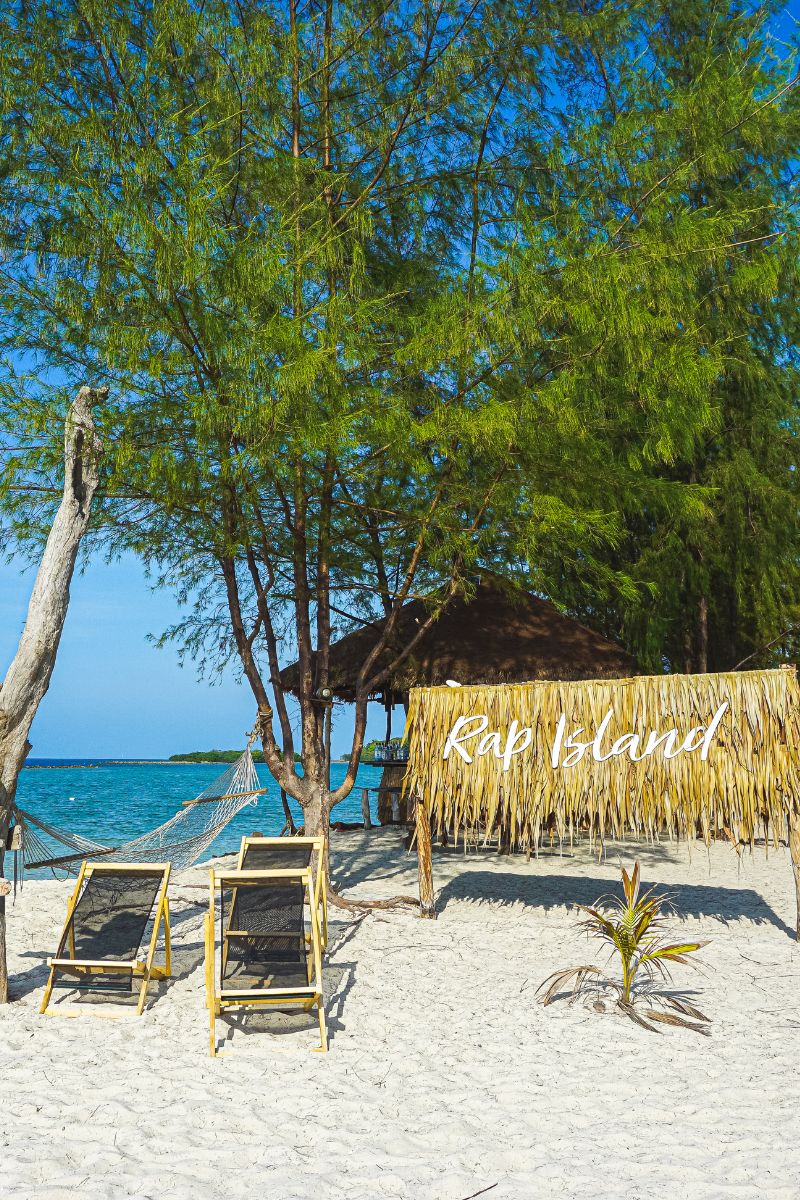 Beach scene with lounge chairs, a thatched hut, and 'Rap Island' sign.