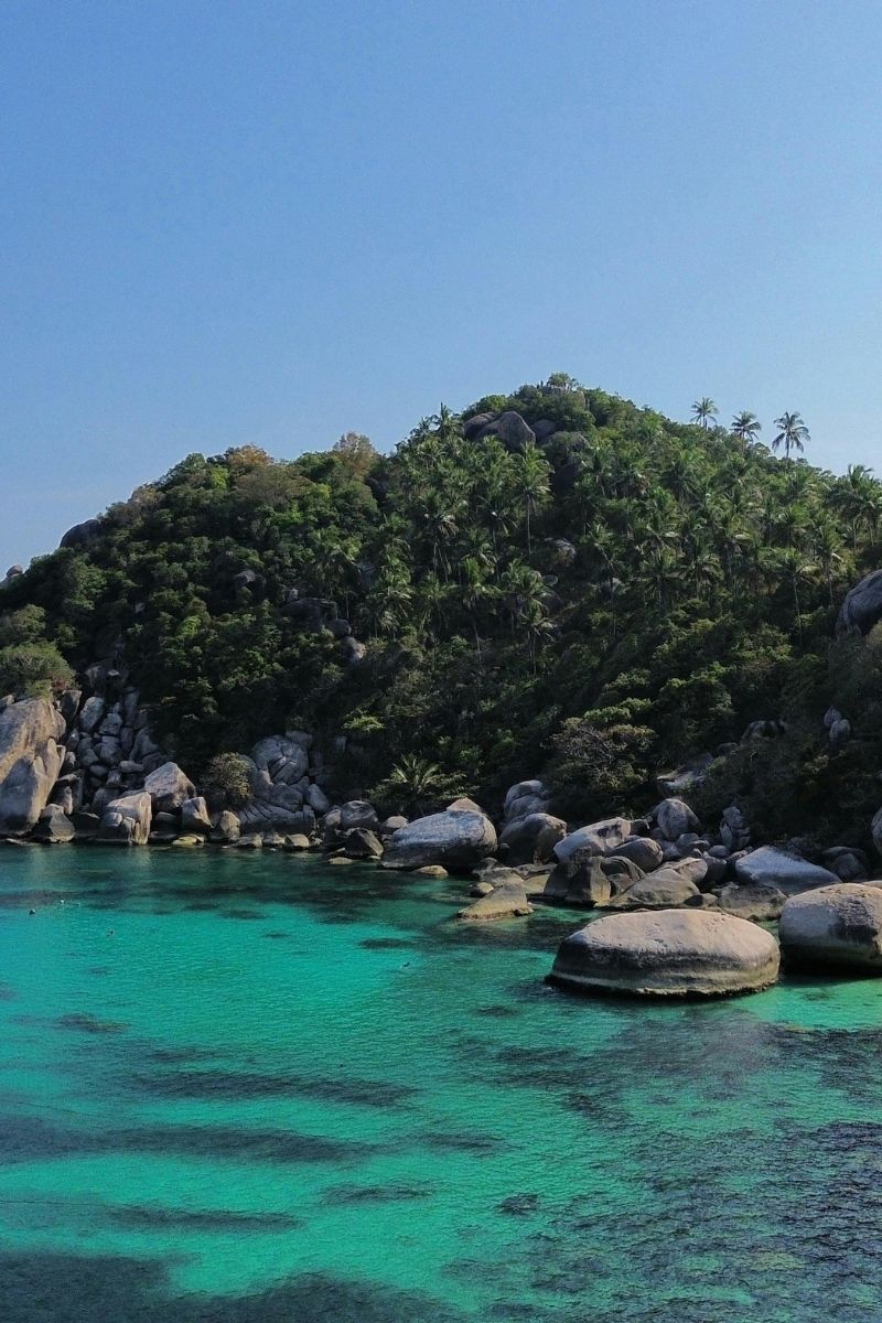 Turquoise waters with a rocky island covered in greenery under a clear blue sky.