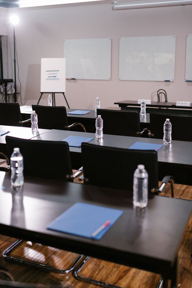 A classroom or conference room setup with rows of black desks and chairs, water bottles on each station, blue folders, and whiteboards on the wall.