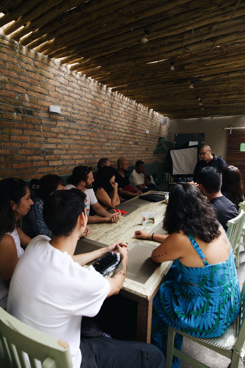 A group of people seated around a long table in a rustic room with exposed brick walls and a bamboo ceiling, with a presenter standing at the front.