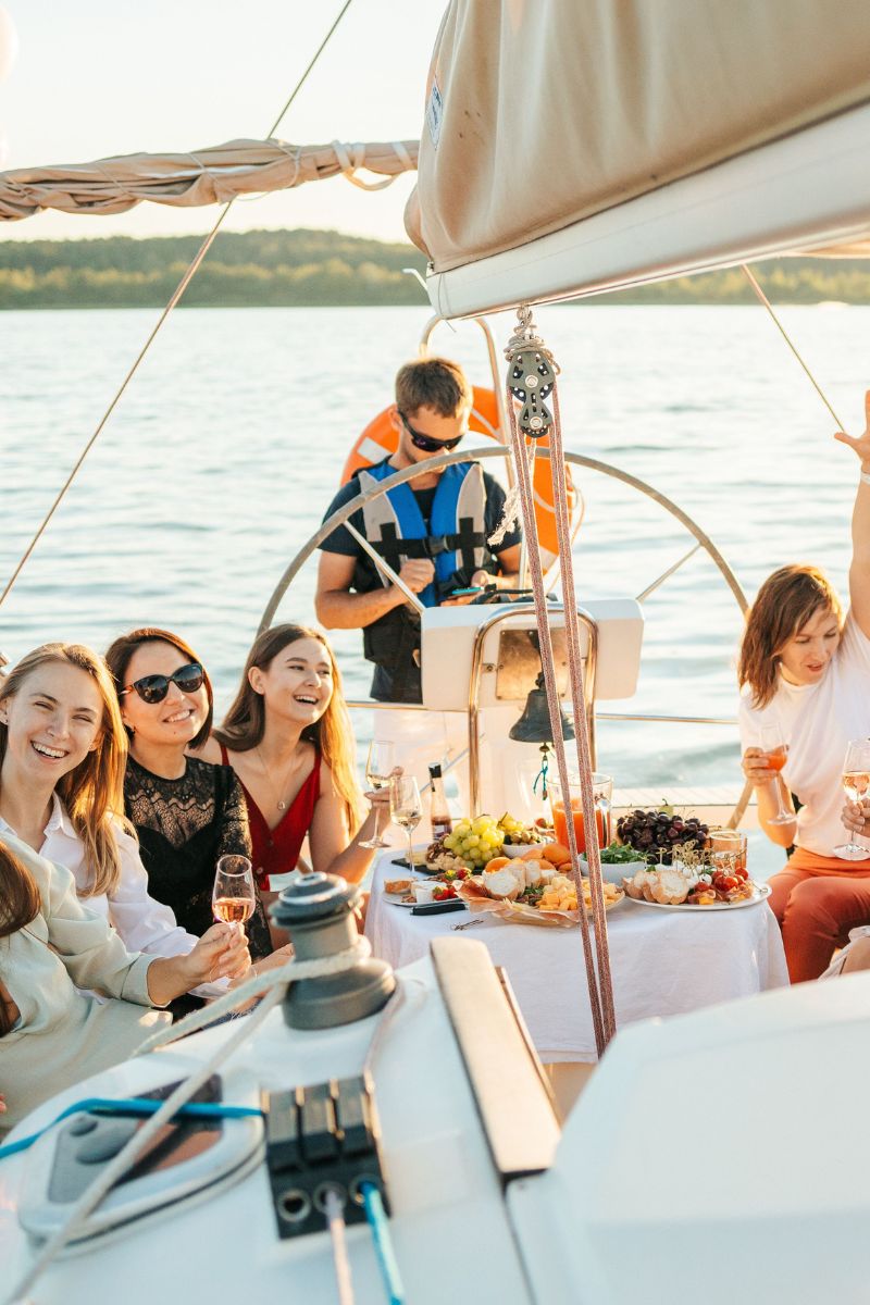 A group of friends enjoying a sunny day on a sailboat with the open water in the background. One person is steering the boat from the helm, while others relax and socialize on deck around a table with food and drinks.