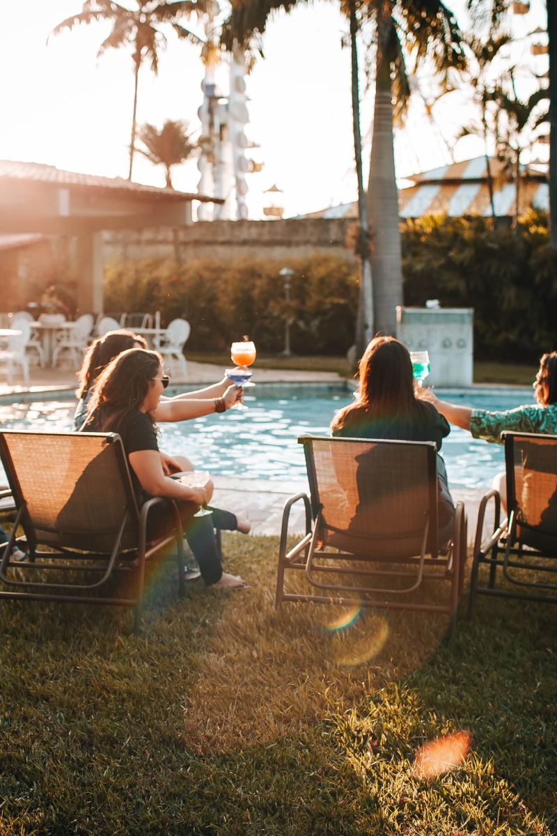 Three people sitting in lounge chairs by a swimming pool at sunset, holding up cocktails. Palm trees, grass, and warm golden light are visible in the background.