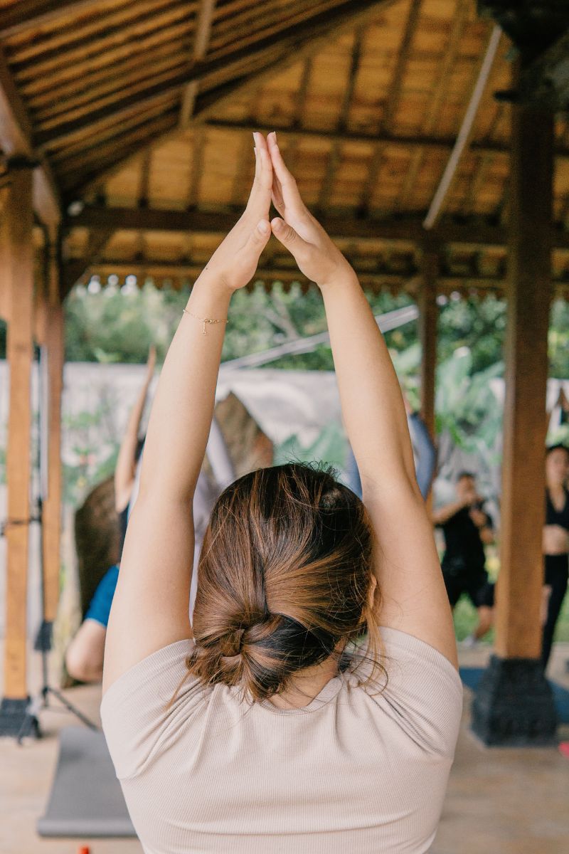 A woman from behind wearing a light beige off-the-shoulder top, performing a yoga pose with her arms raised overhead inside a rustic wooden pavilion.