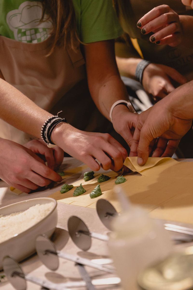 Several people's hands are gathered around a table, making small green dumplings or ravioli on sheets of yellow pasta. A person in a green shirt and beige apron is visible on the left, with other hands and arms reaching in from the right.