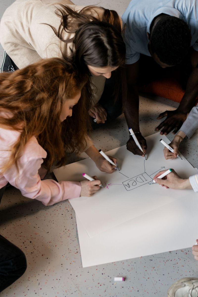 Several people gathered around a large white sheet of paper on the floor, writing and drawing with markers. The scene is viewed from above, showing a collaborative workspace with a speckled gray floor.