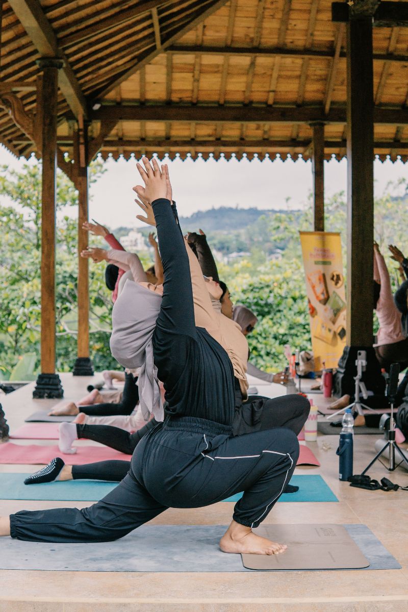 A group of people practicing yoga outdoors under a wooden pavilion, with one person in the foreground performing a deep lunge pose with arms raised overhead. The setting features green hills in the background, and other participants are visible on yoga mats.
