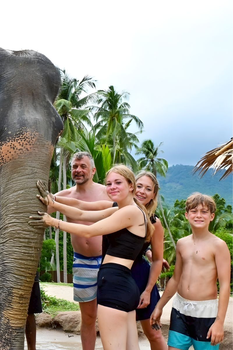 Family posing with an elephant in a tropical setting with palm trees and mountains.