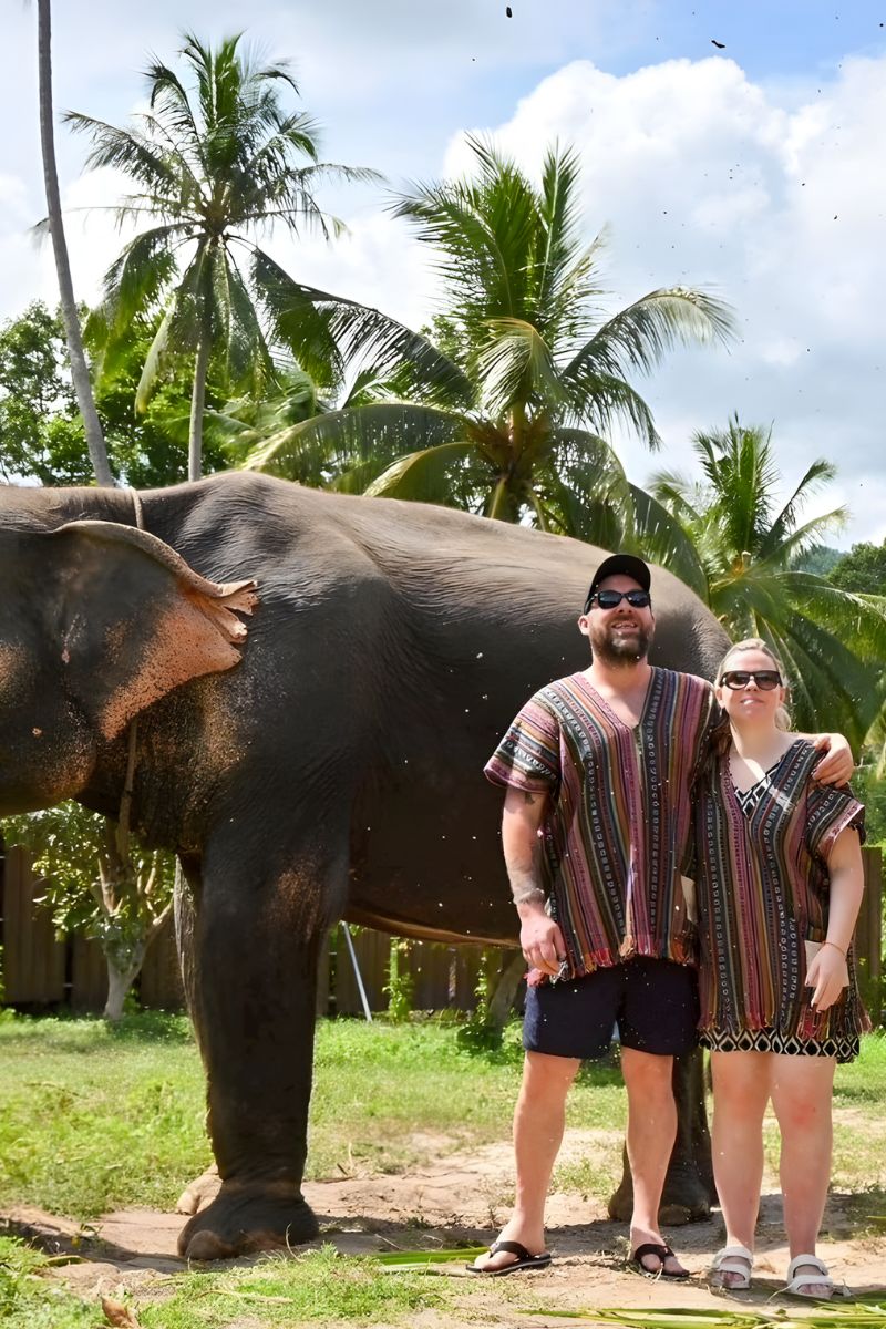Two people in matching outfits standing next to an elephant with palm trees in the background