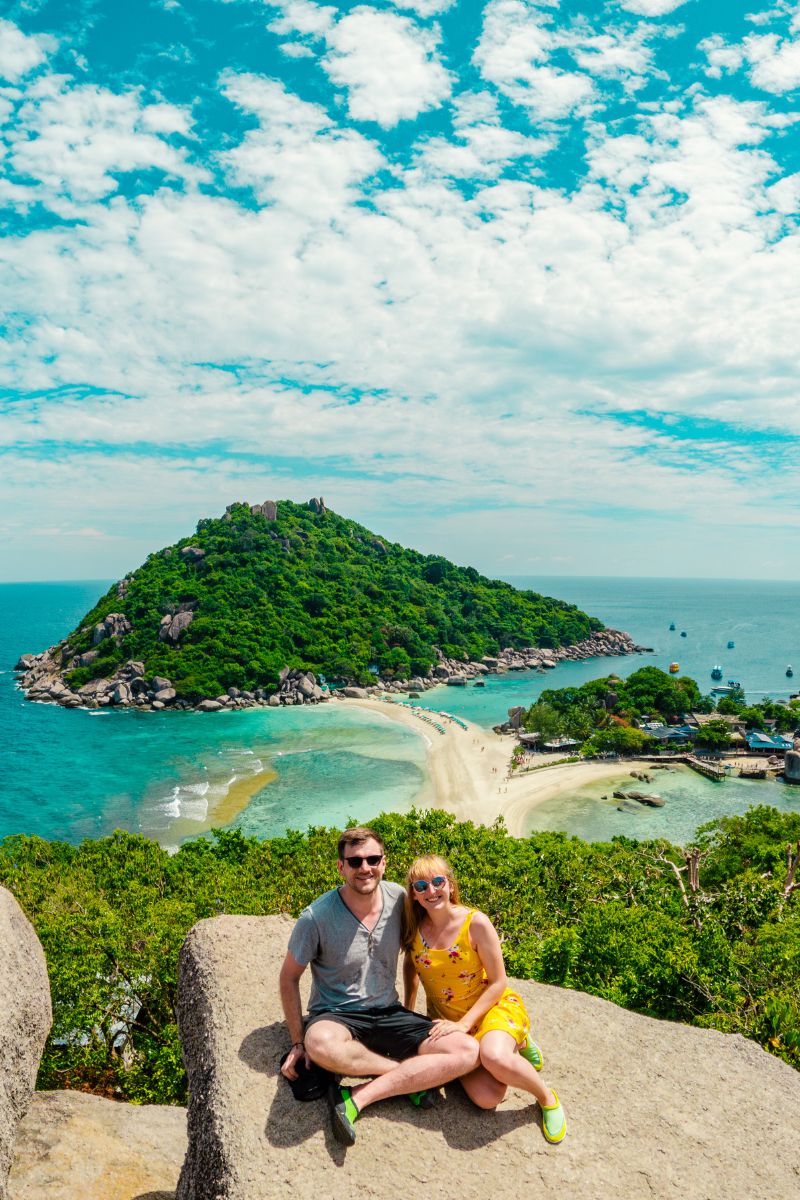 Two people sitting on a rock overlooking a tropical island with clear blue water and greenery.