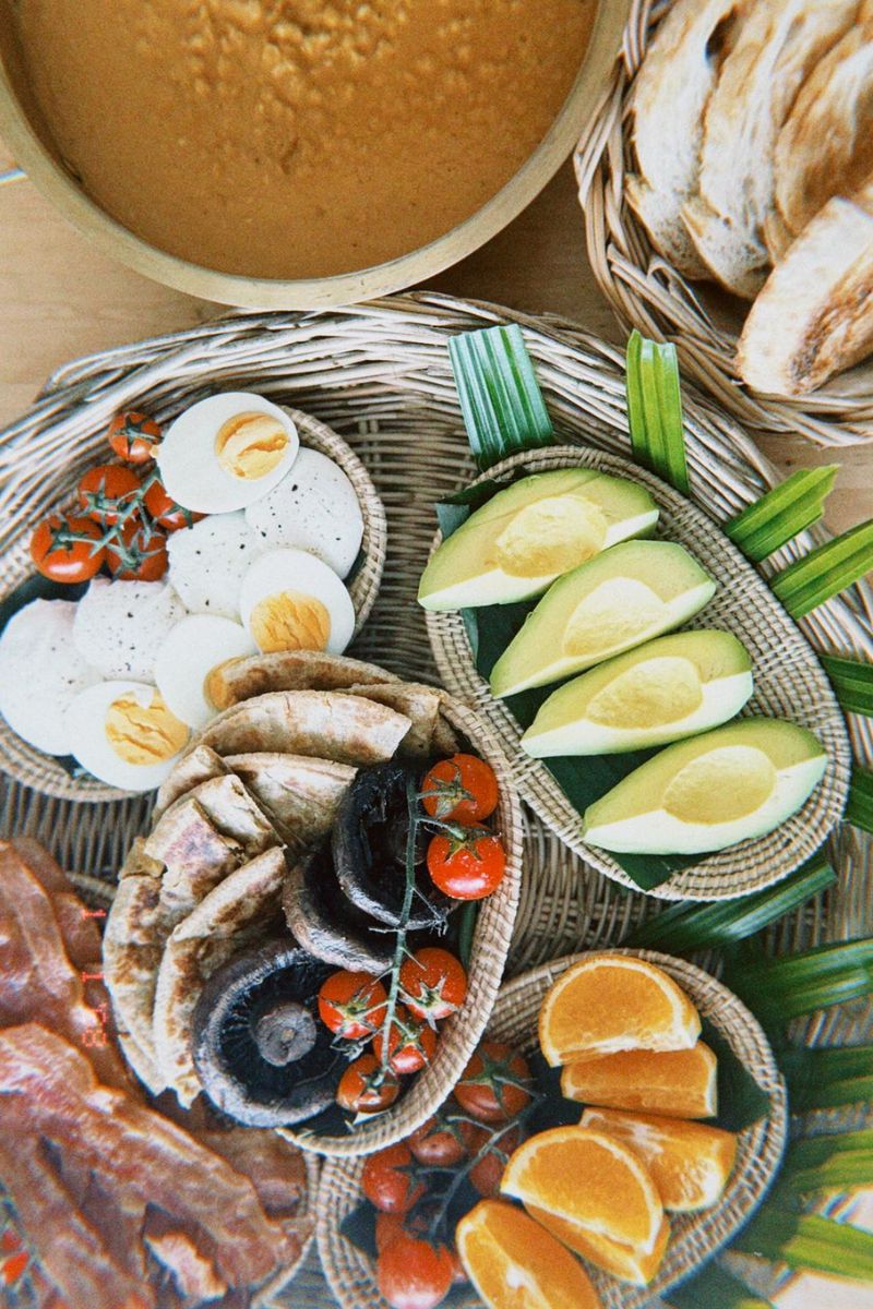 Assorted food items including eggs, avocados, tomatoes, oranges, and bread on a woven mat.