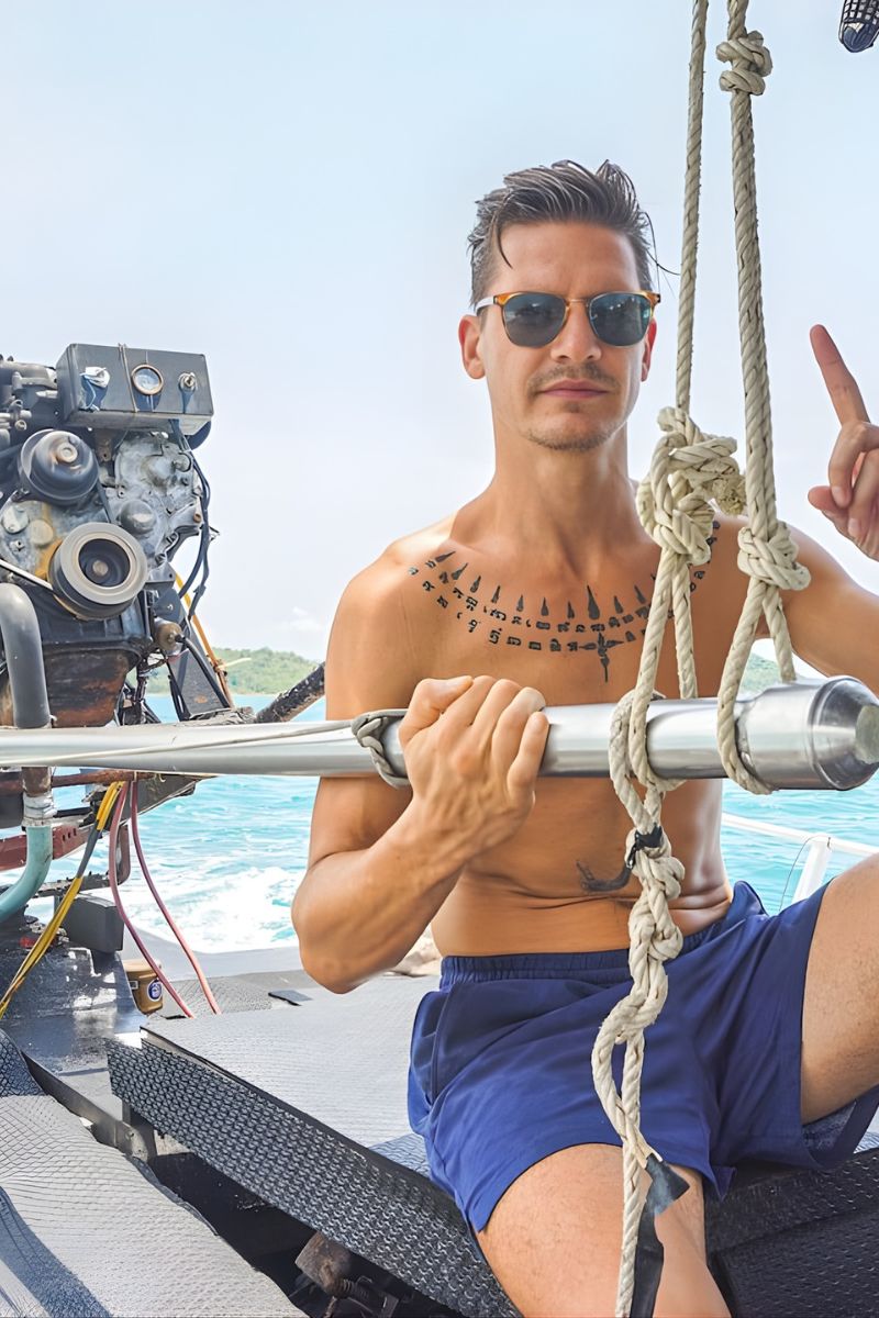 Man on a boat wearing sunglasses and blue shorts, with a scenic background.