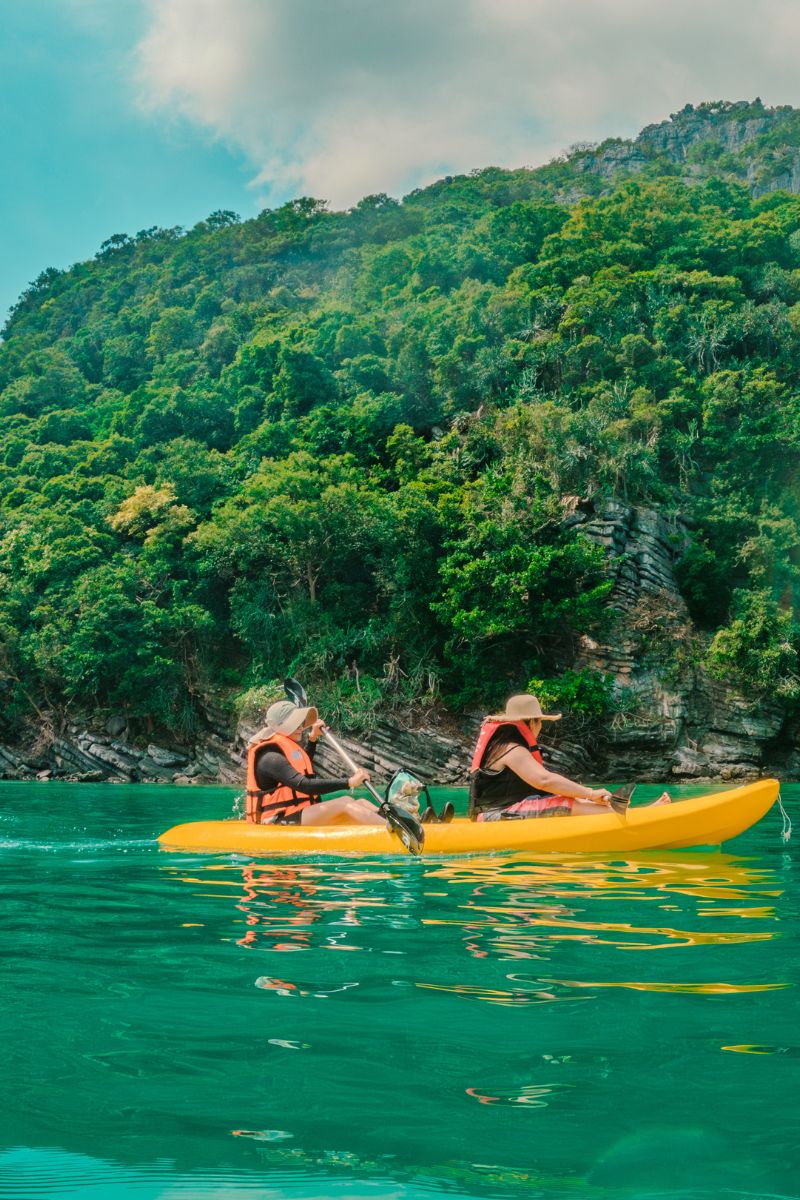 Two people in a yellow kayak on a clear lake with a forested hillside in the background.