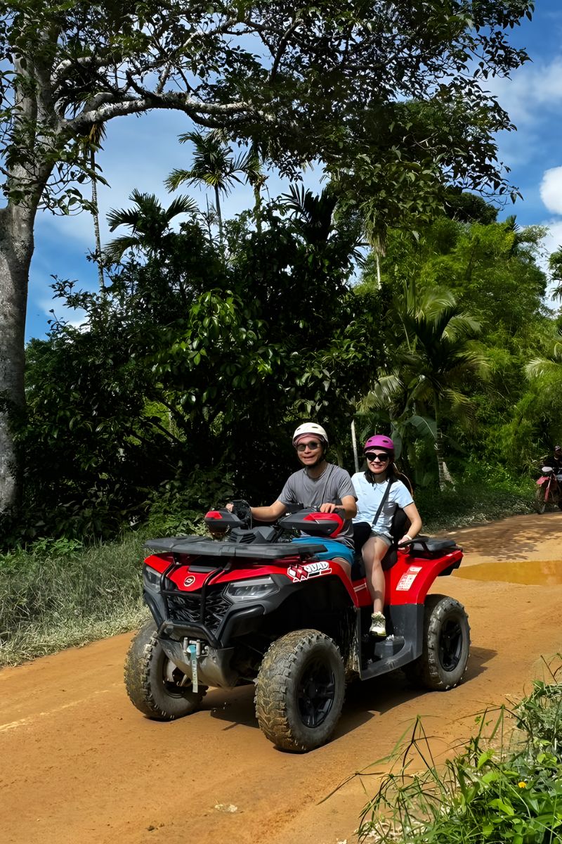 Two people riding a red ATV on a dirt road surrounded by greenery