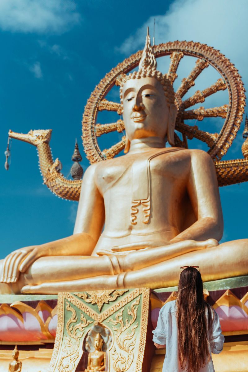 Gold Buddha statue with a person in a white dress standing in front, against a blue sky.