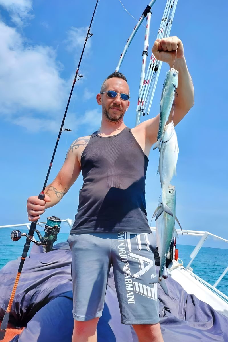 Man on a boat holding a fish and a fishing rod with a clear blue sky in the background