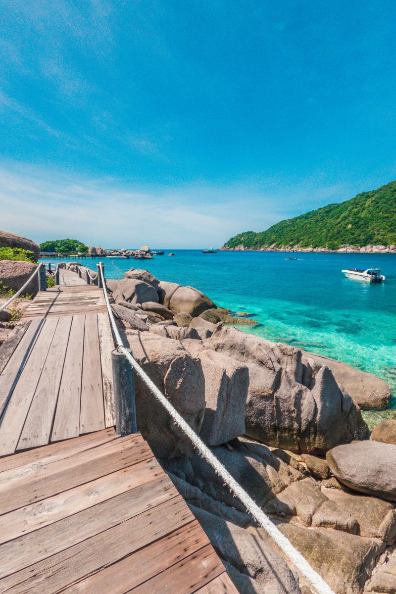 Wooden dock extending into clear blue water with rocky outcrops and a boat in the distance.
