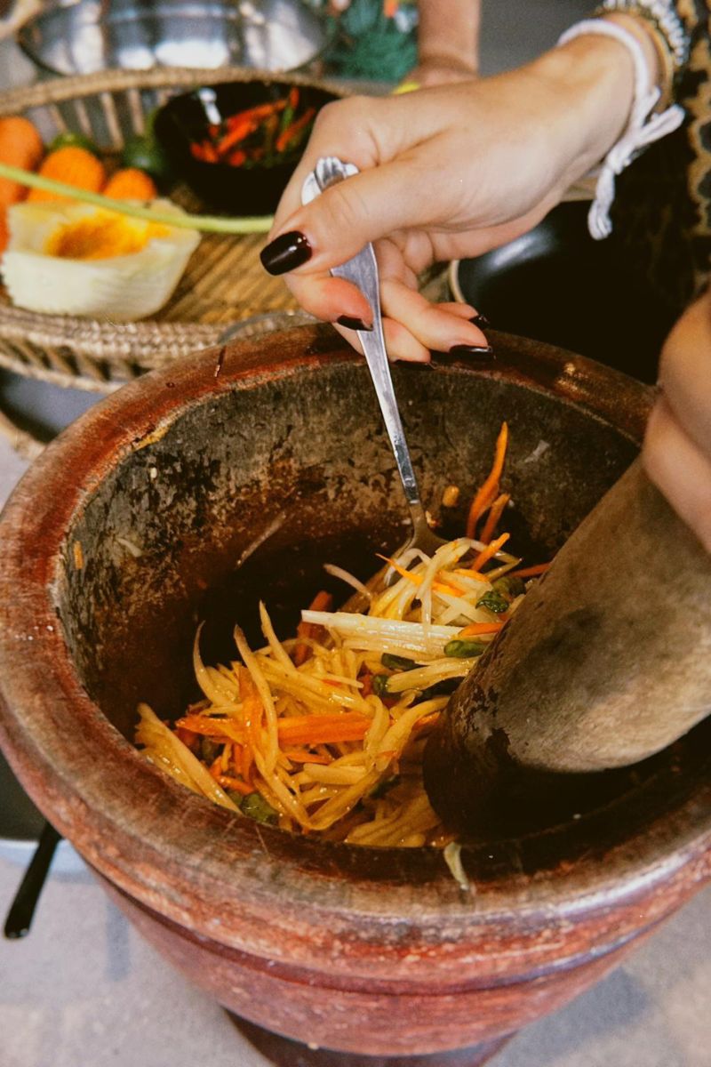 Person using a pestle to grind ingredients in a mortar with a blurred background of fruits and vegetables.