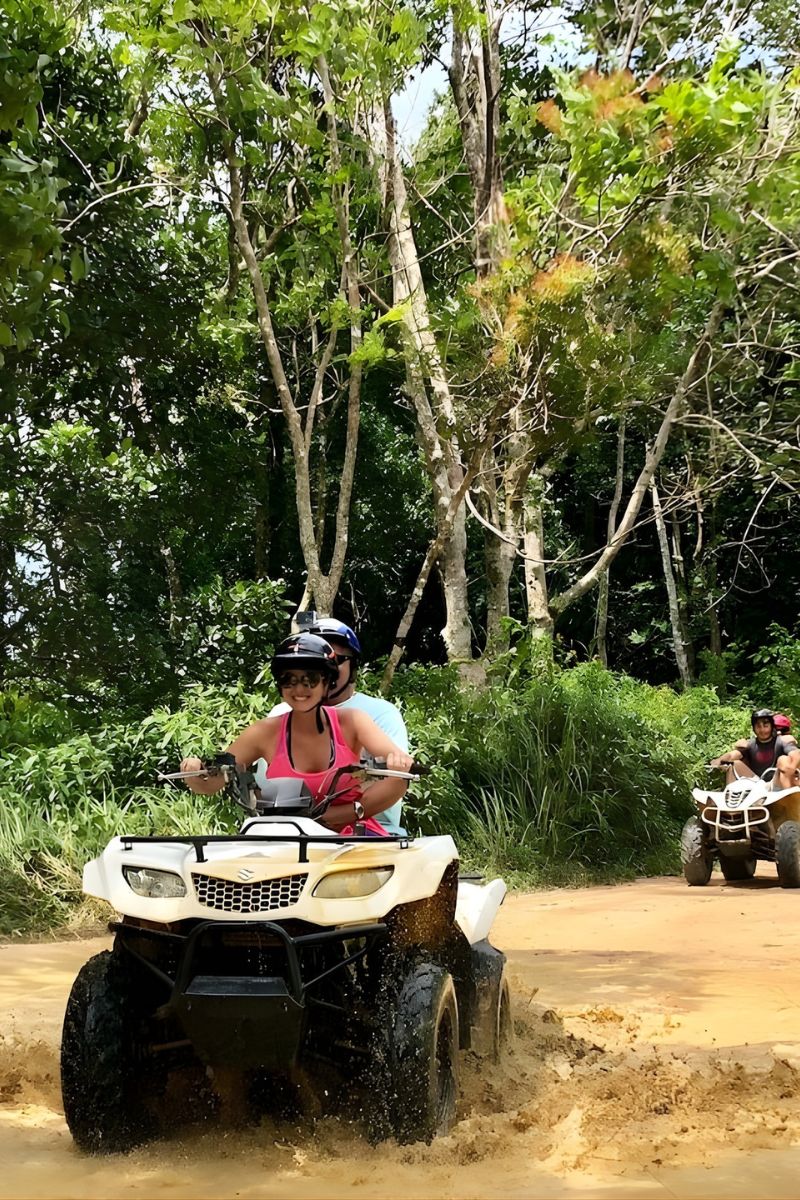Two people riding ATVs on a dirt road surrounded by trees