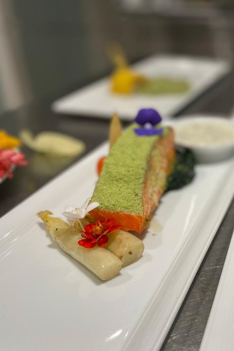 Delicate dessert with green crumbly top, red and white floral garnish, and small white flowers on a white plate.