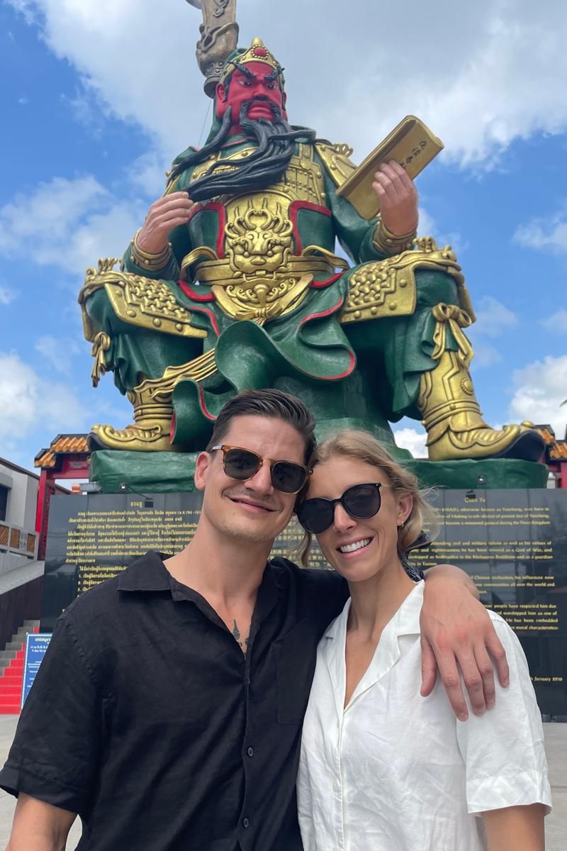Two people posing in front of a large statue with a blue sky background