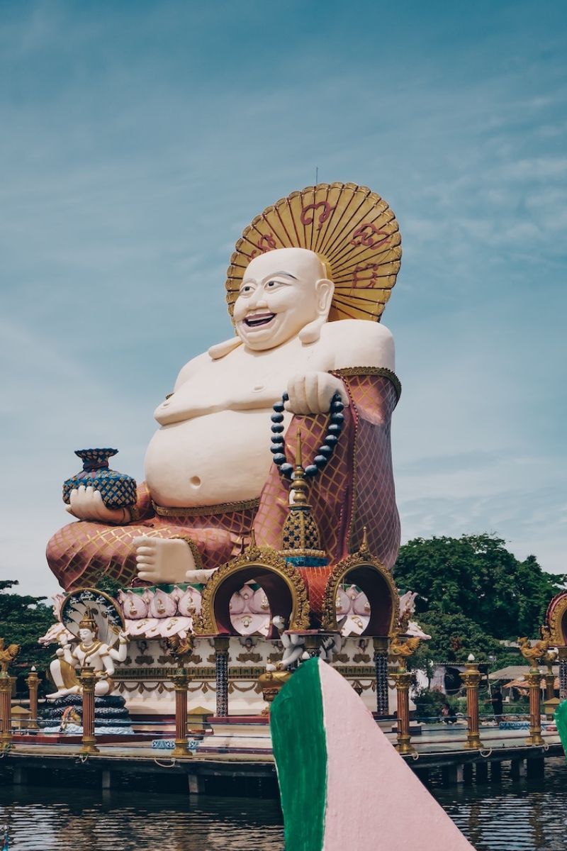 Large statue of a laughing Buddha with a decorative umbrella against a blue sky.