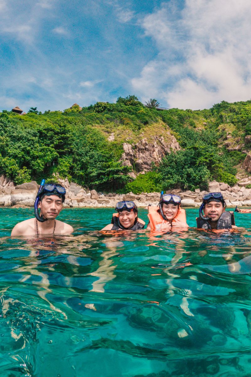 Four people snorkeling in clear blue water with a scenic background of greenery and rocks.